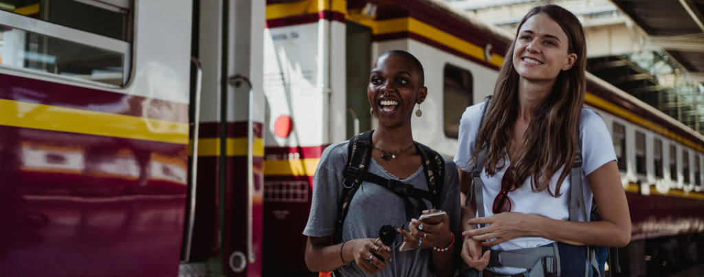 Deux jeunes filles qui sont sur un quai, devant un train et qui sont souriantes